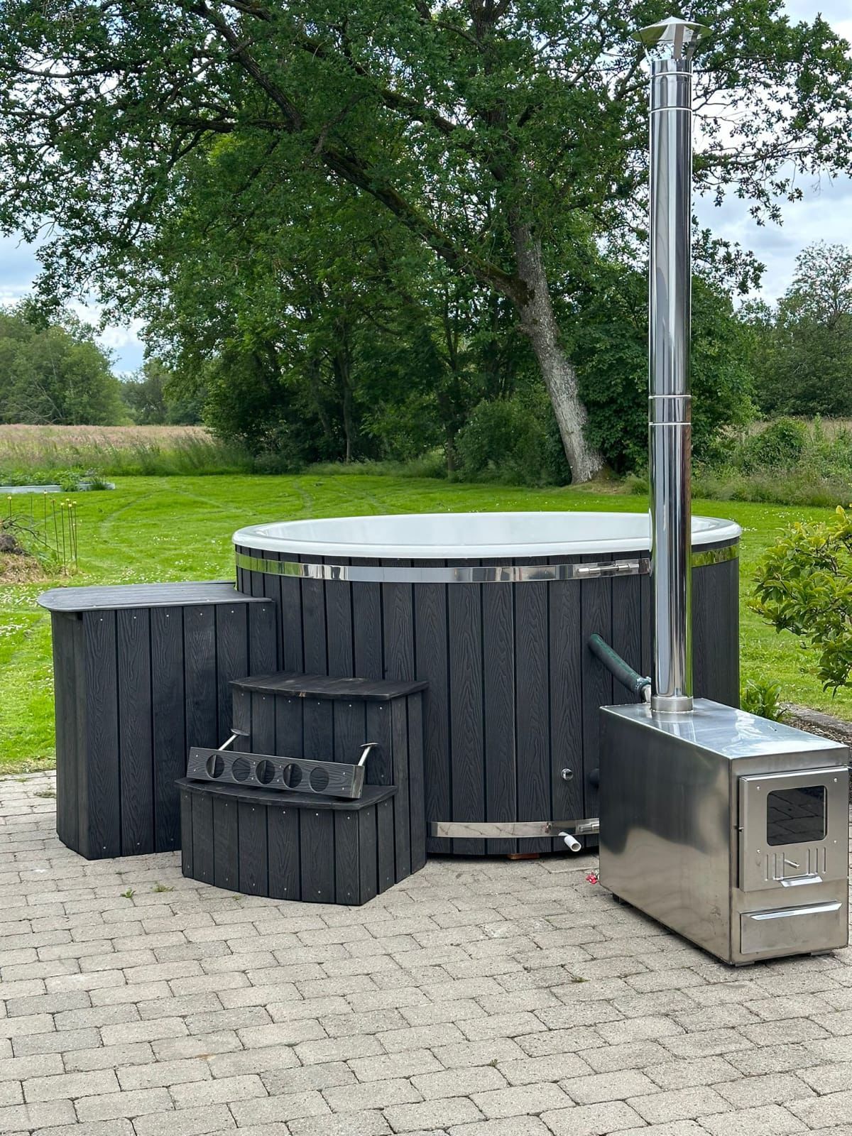 Round outdoor hot tub with dark WPC cladding and external stainless steel heater